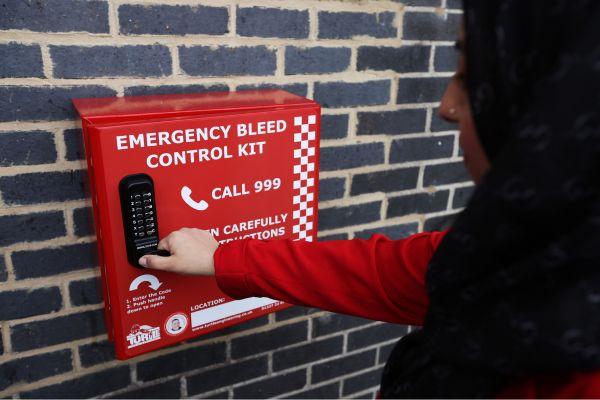 A red bleed kit box, attached to a dark brick wall, with a woman reaching towards it.